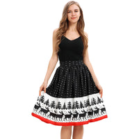 A young woman with long blonde hair is wearing a sleeveless black top paired with a knee-length, high-waisted skirt featuring a festive Christmas print. The skirt has white polka dots and a scenic pattern of deer and trees at the hem, bordered by a red stripe. She smiles slightly and stands against a plain white background.
