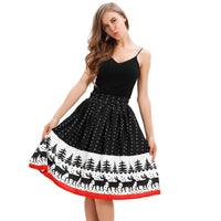 A woman with long, wavy hair poses playfully, wearing a sleeveless black top and a knee-length Christmas print skirt. The skirt is high-waisted and features white polka dots with a white band at the bottom adorned with black silhouettes of reindeer and pine trees, accented by a red hemline.